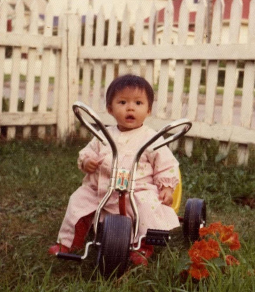 Angel as a toddler on a tricycle in front of a white picket fence