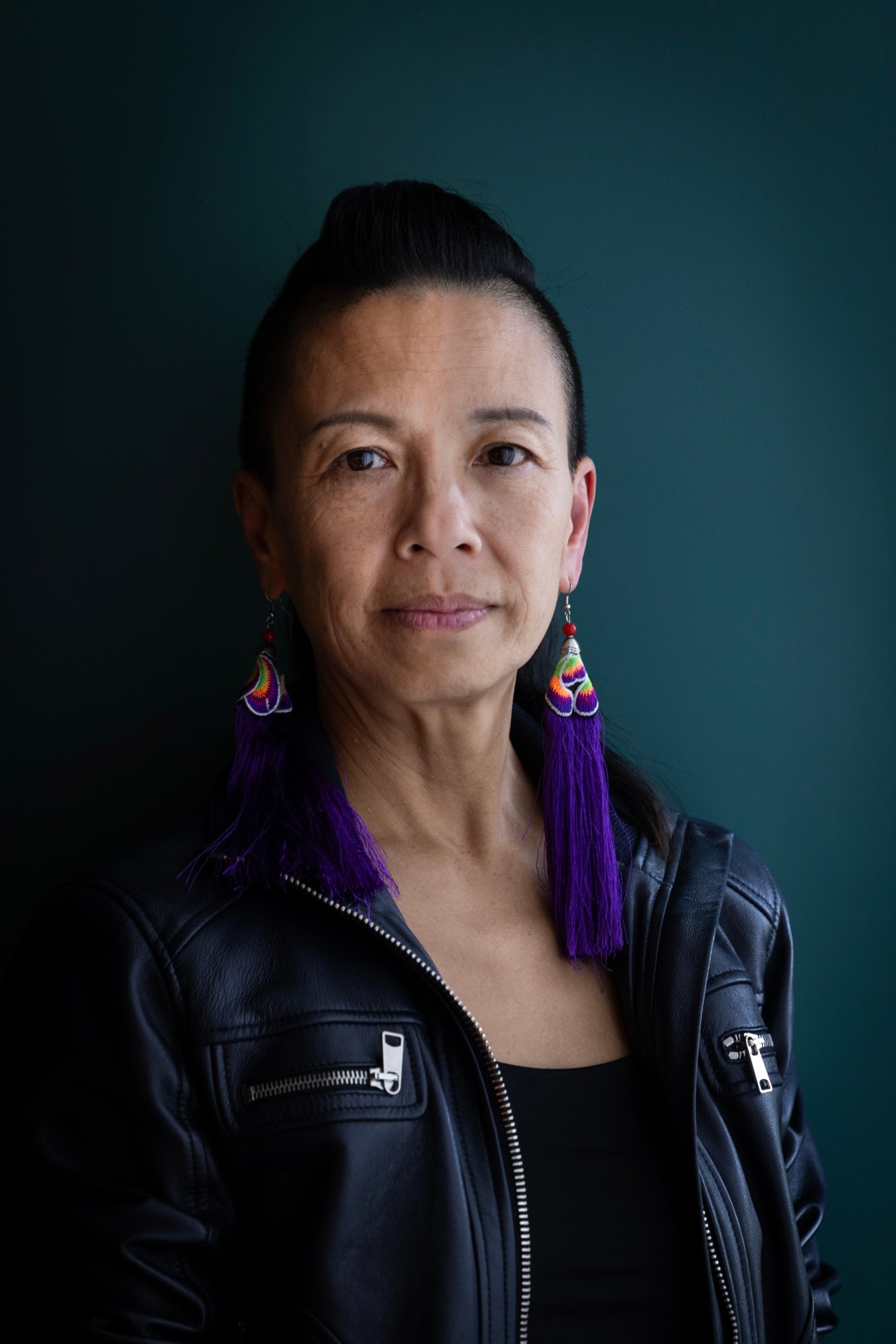 Angel Cheng in a black leather jacket and purple tassel earrings against a deep teal wall
