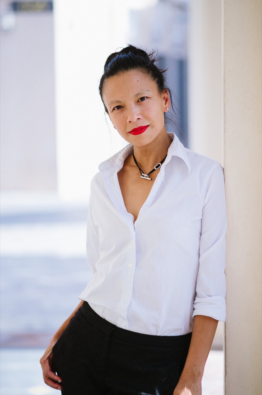 Angel Cheng in a white shirt and red lipstick, leaning against a pale column