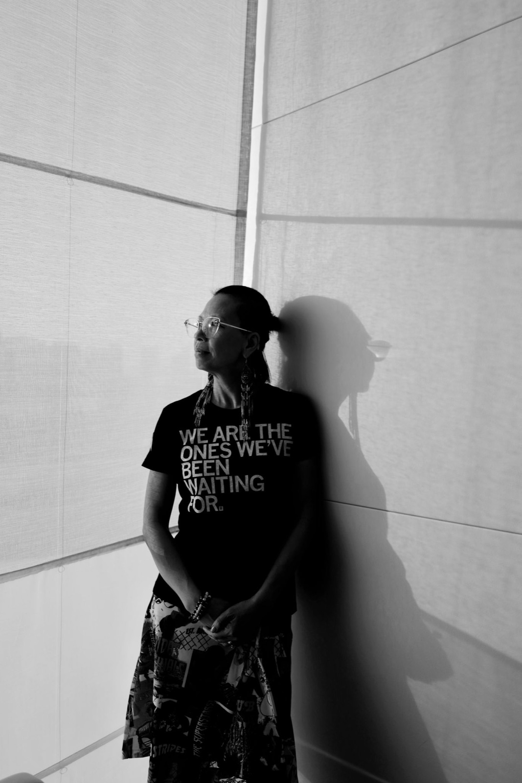 Black and white photo of Angel Cheng leaning against a sunlit wall in a T-shirt that reads We are the ones we've been waiting for