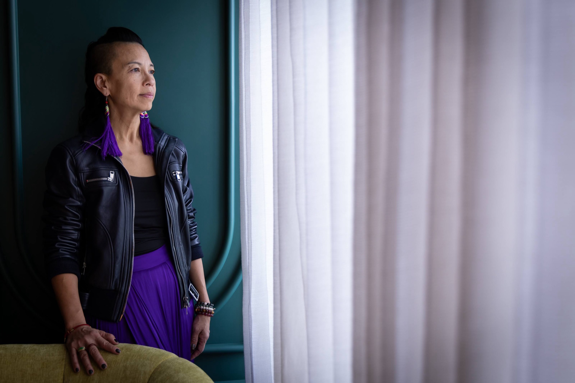 Angel Cheng in a leather jacket and violet skirt, standing by a sheer curtain in a teal room